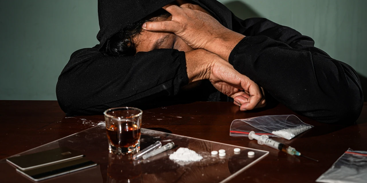 A person sits at a table surrounded by drugs and alcohol, illustrating the destructive impact and emotional burden of drugs addiction.