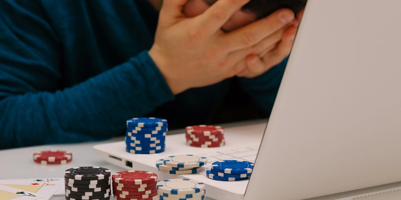 A person sits with their head in their hands beside poker chips and a laptop, showing the emotional stress and consequences of gambling addiction.