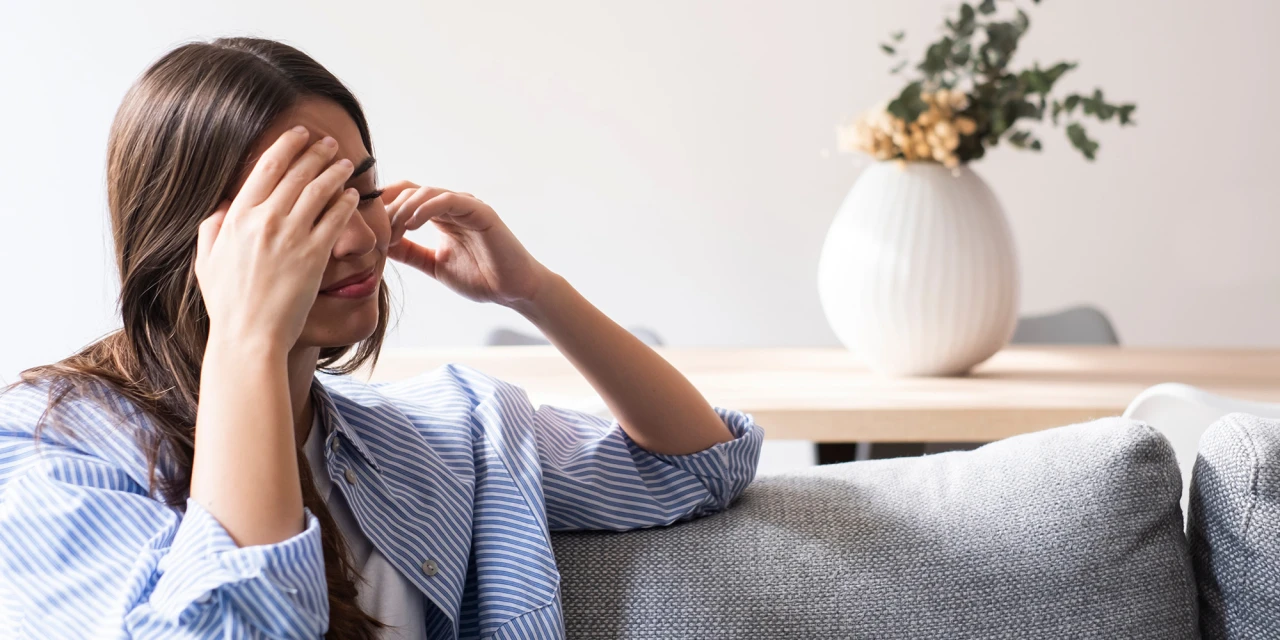 A woman sits on a sofa holding her head, showing physical tension, mental exhaustion and the impact prolonged stress can have on your wellbeing.