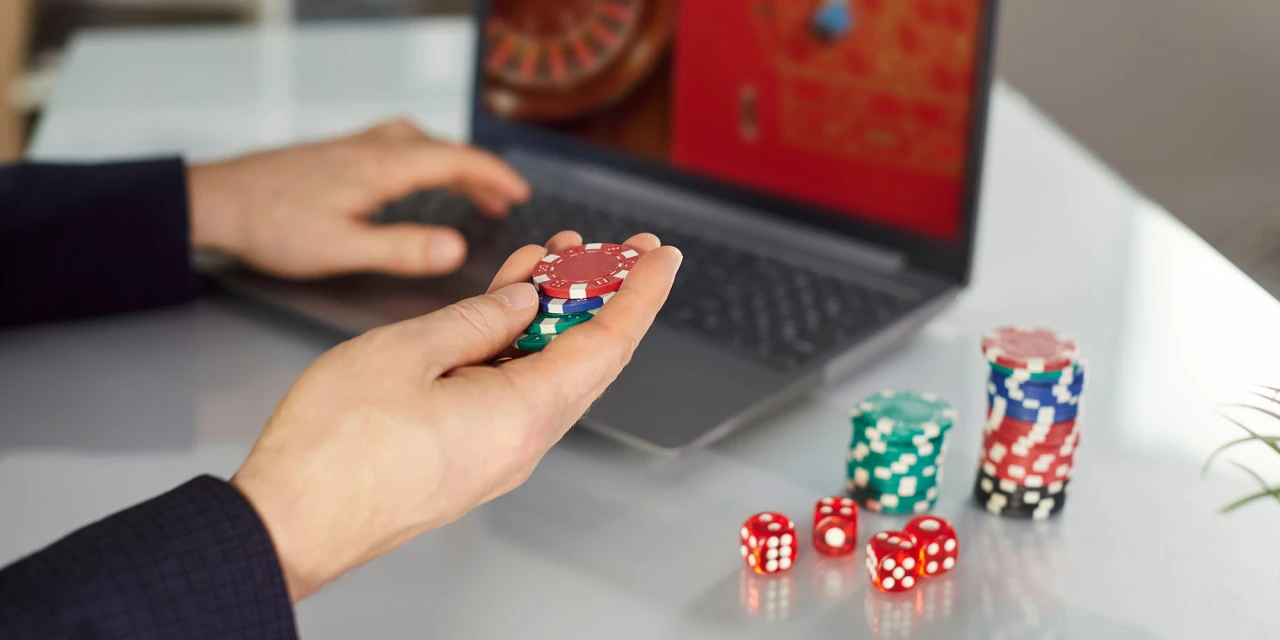 A person holds poker chips while gambling on a laptop, illustrating the accessibility and financial risks connected to gambling addiction.