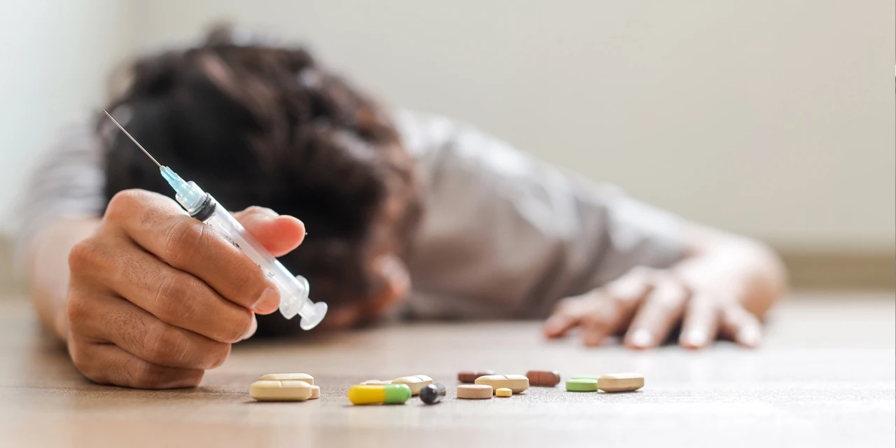 A person lies on the floor holding a syringe near scattered pills, showing the physical and emotional impact of heroin addiction.