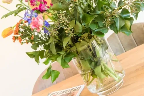 Bos bloemen op een houten tafel, met op de achtergrond een witte muur en lichtgekleurde stoelen.