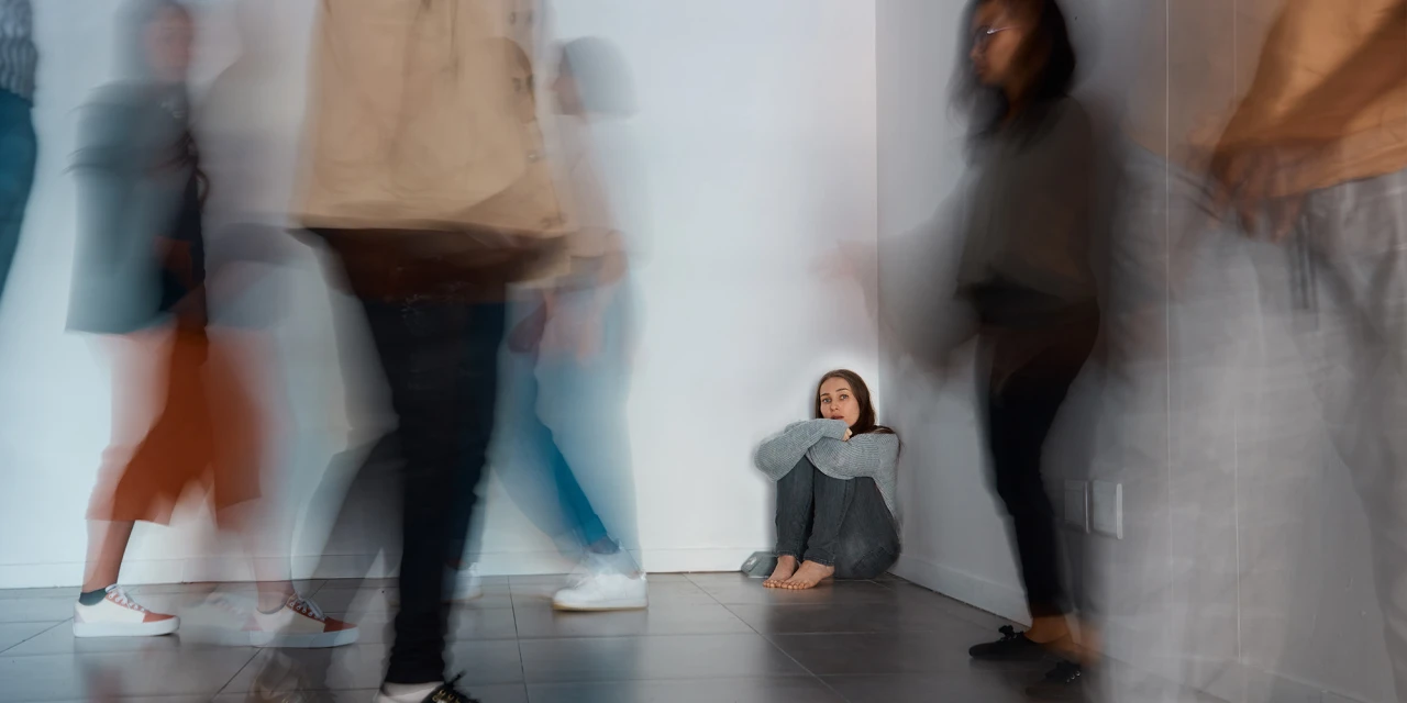 A young woman sits alone against a wall while blurred people move around her, showing isolation, fear and the emotional impact of panic attacks.