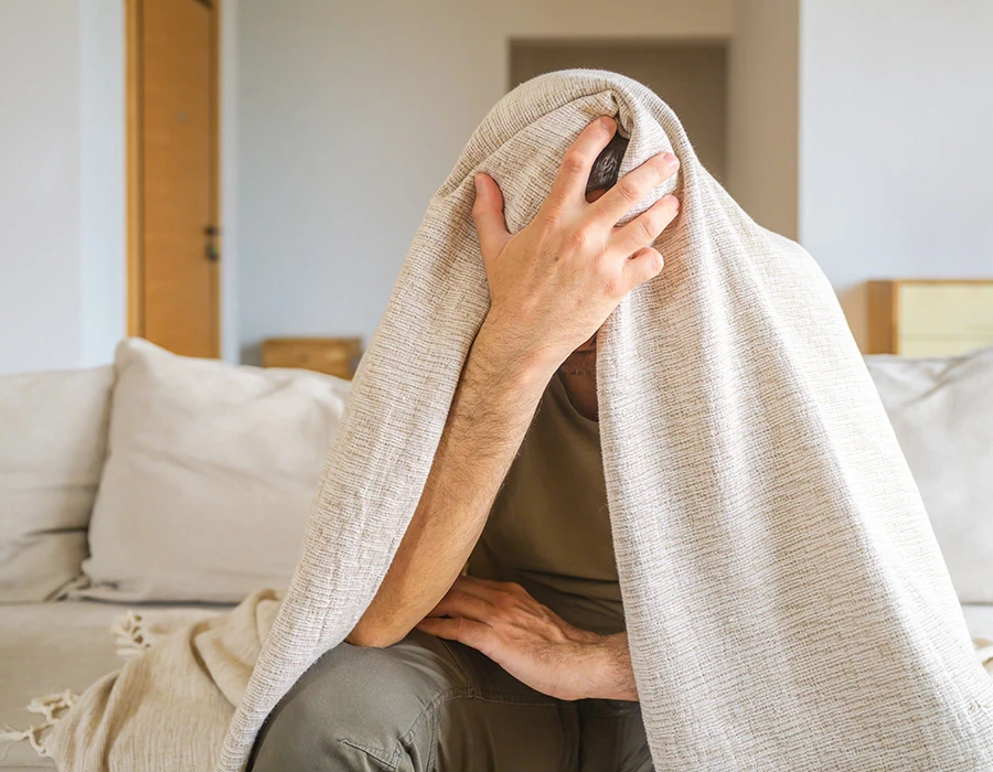Man covering his head with a blanket while experiencing possible alcohol-related symptoms.