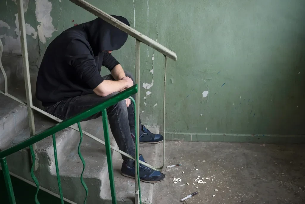 A man in a black hoodie and jeans sitting on a staircase, with his arms resting on his knees and his head bent.