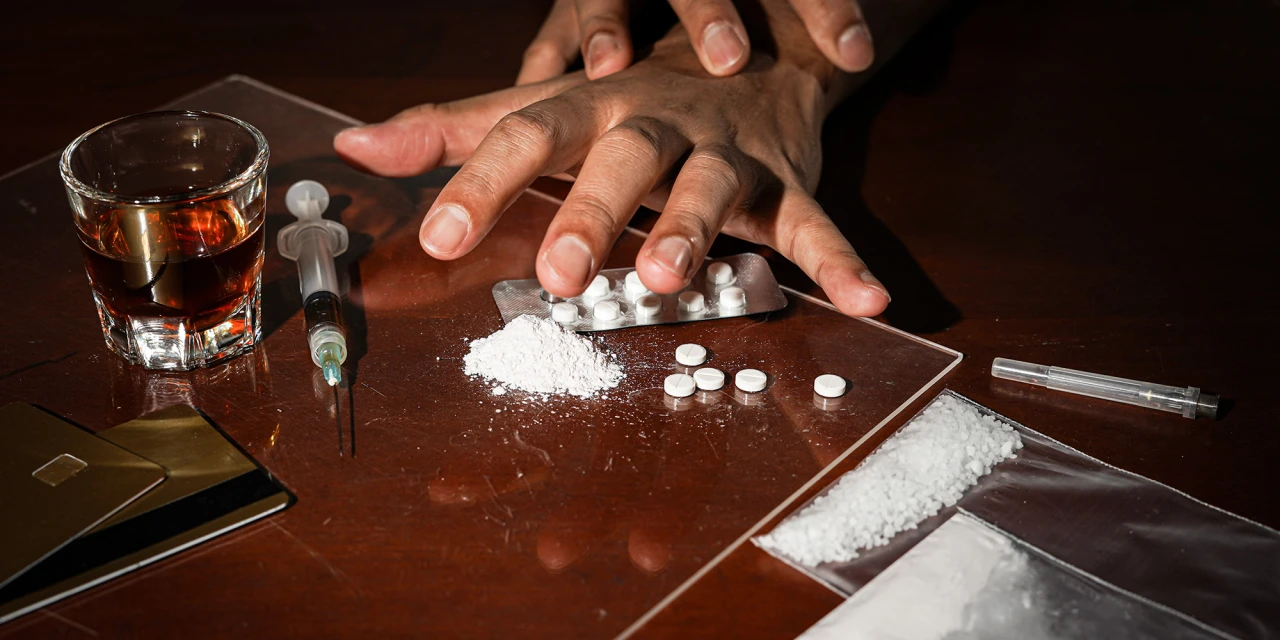 A person reaches towards white powder, pills, alcohol and syringes on a table, illustrating the dangerous reality and health risks linked to heroin addiction.
