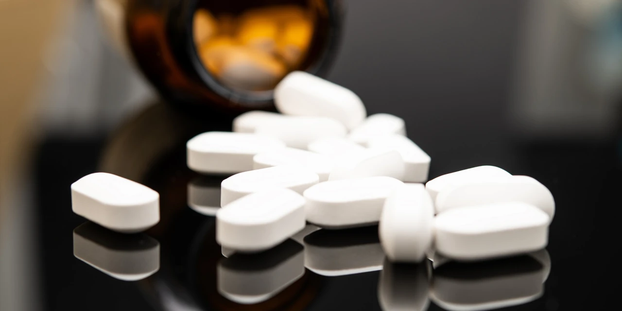 Several white opioid pills lie on a reflective surface beside an open medicine bottle, symbolising prescription drug misuse and opiod addiction.