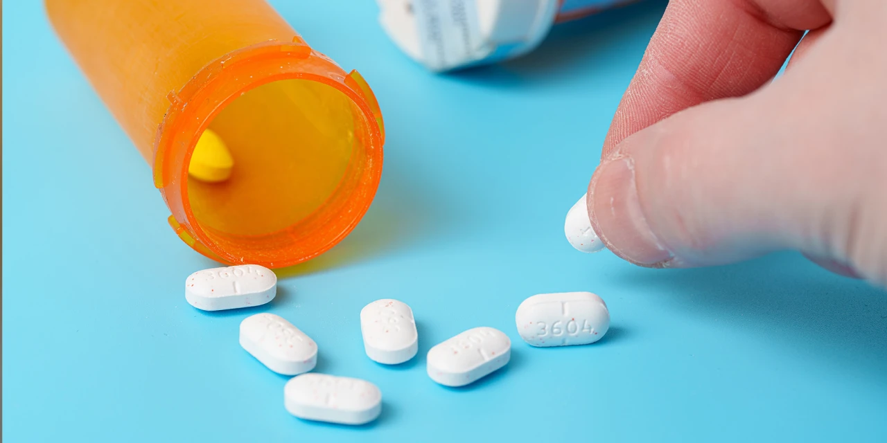 White opioid tablets spill from a prescription bottle while a hand reaches for more medication, representing patterns associated with opiod addiction.