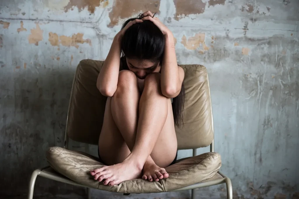 A woman hugging herself and holding her head, sitting on a chair with her legs folded against an industrial wall.