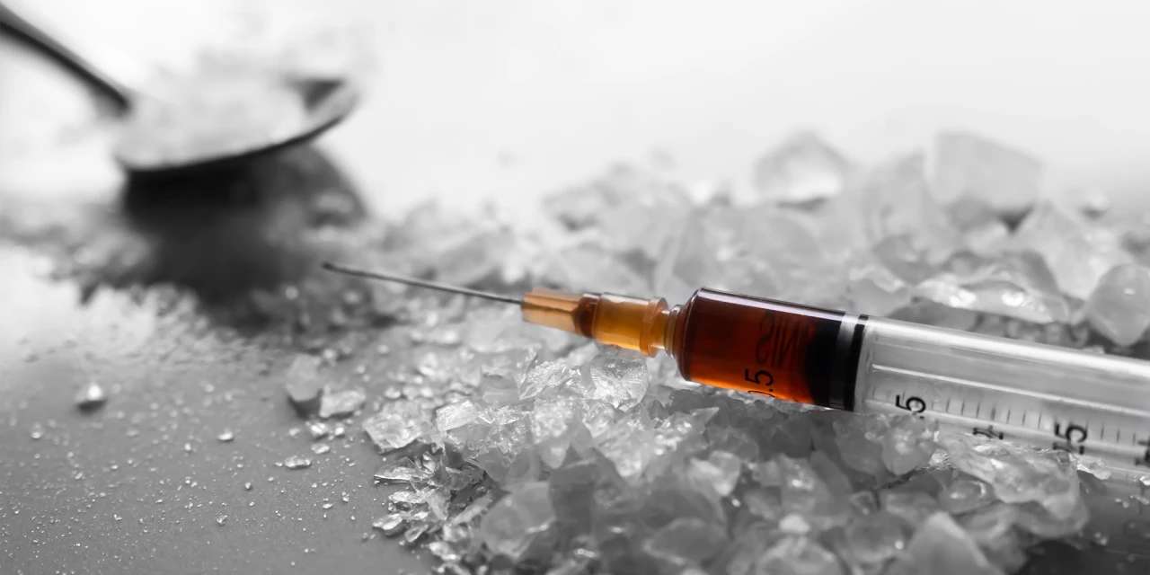 A syringe lies beside crystal-like substances and white powder, showing drug paraphernalia often associated with amphetamine addiction and substance misuse.