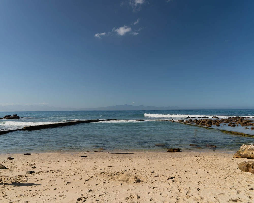 A tidal pool by the sea, with rocks, a sandy beach and mountains on the horizon.