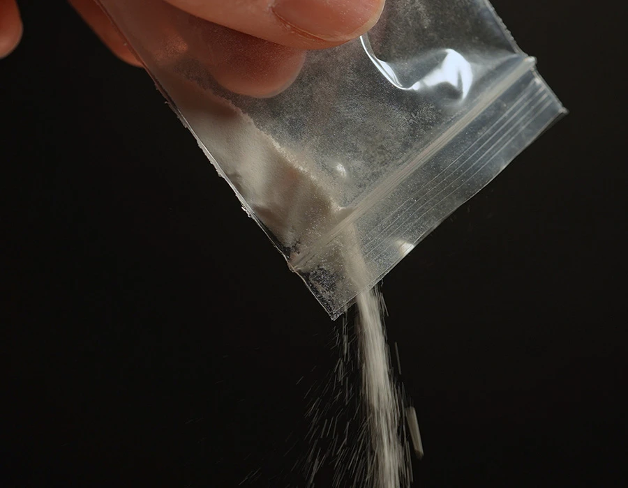 White ketamine powder pouring from a small plastic bag held between fingers, dark background