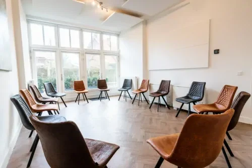 Behandelkamer met witte muren, een grote raampartij en bruine leren stoelen in een kringopstelling.