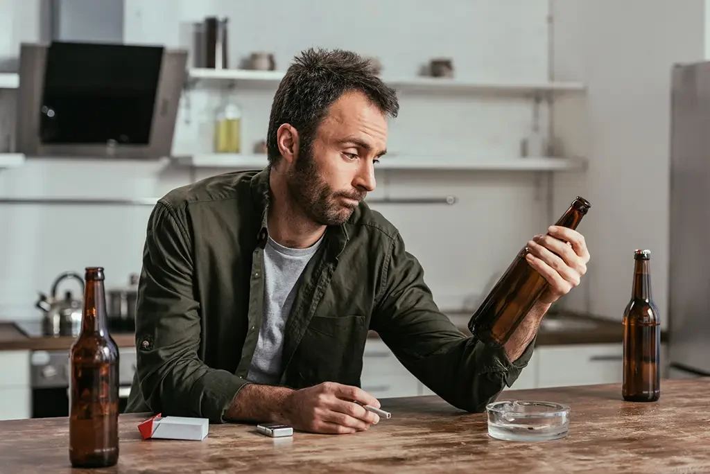 A man sits at a kitchen table surrounded by empty bottles. He gazes doubtfully at a bottle in his hand.