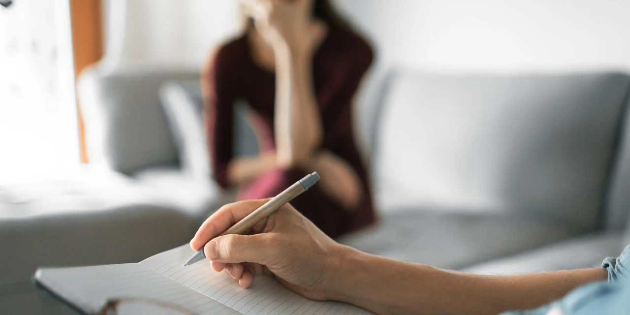 A therapist takes notes while a woman sits in the background during a conversation about trauma, emotional distress and PTSD symptoms.