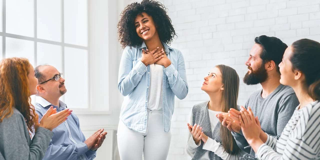 A woman stands with a smile and her hands over her heart as a diverse group of people sits in a circle and claps for her.