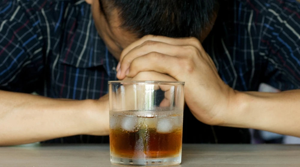 Person experiencing alcohol withdrawal symptoms, sitting with hands clasped beside a drink.