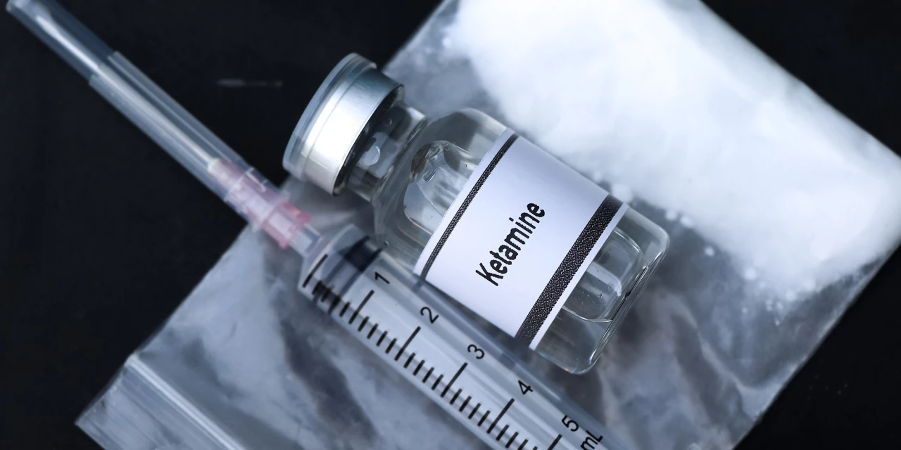 A ketamine vial and syringe rest on a plastic bag filled with white powder, representing the harmful effects and serious consequences of Ketamine addiction.
