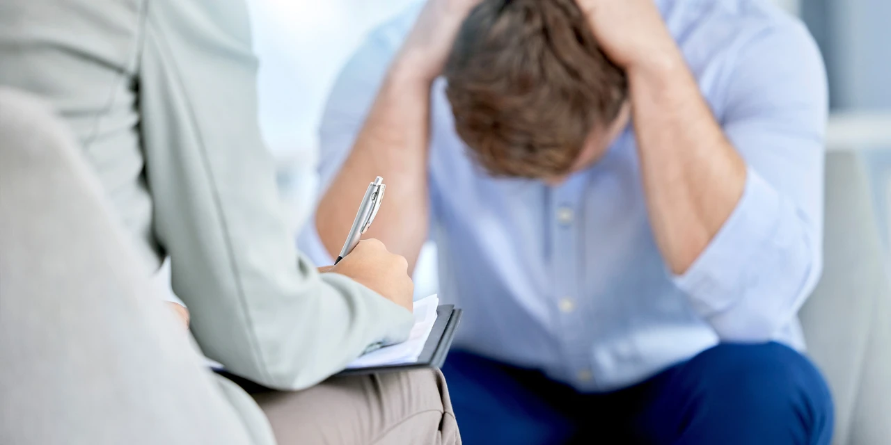 A man sits with his head in his hands during a therapy session, showing emotional exhaustion and stress related to burn out.