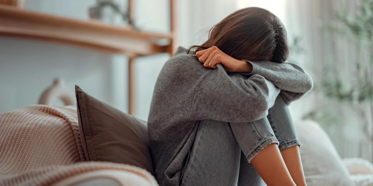 A woman sits curled up on a sofa with her head resting on her arms, reflecting sadness, exhaustion and the emotional weight of depression.