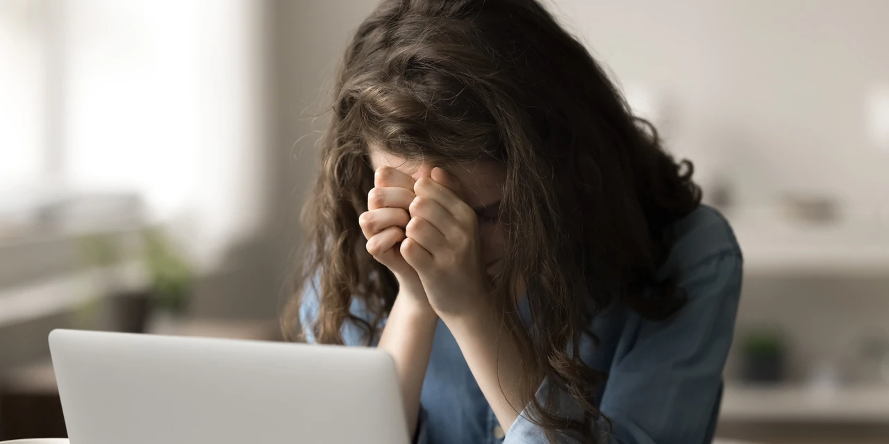 A woman sits behind a laptop with her face in her hands, reflecting emotional exhaustion, pressure and the effect stress can have on daily life.