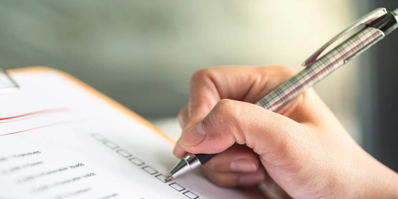 A close-up of a hand using a pen to check boxes on a formal medical document attached to a clipboard.